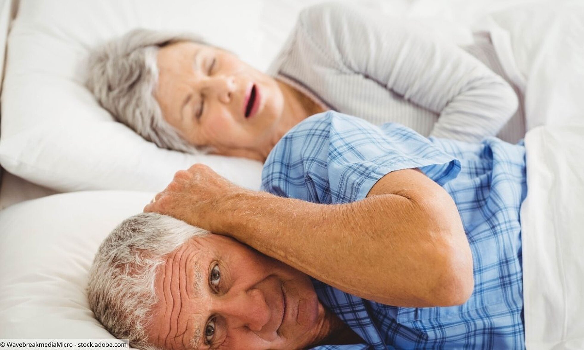 Älteres Ehepaar liegt zusammen im Bett, er hält sich die Ohren zu.