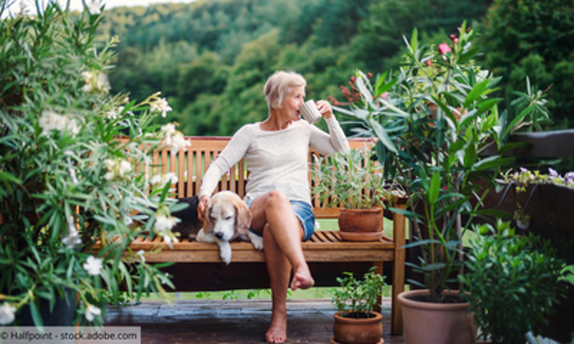 Ältere Frau sitz mit Hund im Garten und trinkt Kaffee