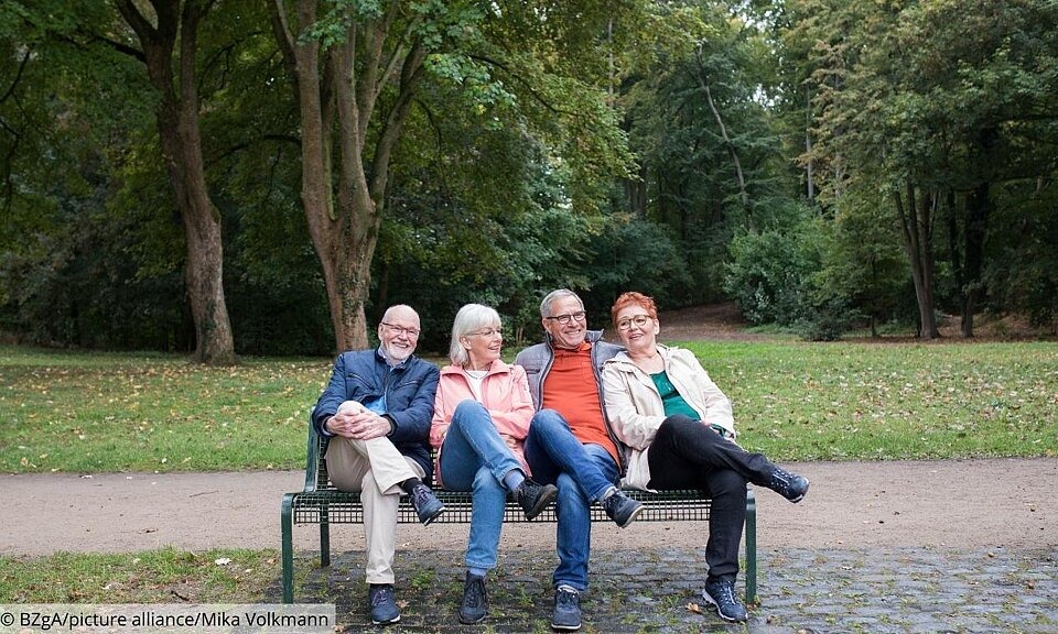 4 ältere Menschen sitzen auf einer Parkbank mit übereinandergeschlagenen Beinen.