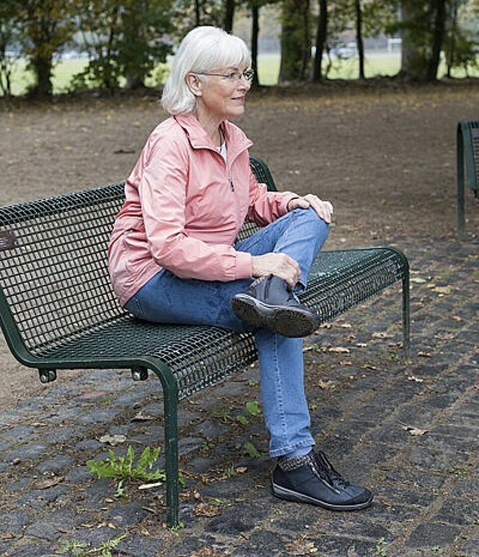 Frau sitzt auf Parkbank mit linkem Bein angewinkelt auf rechtem Bein.