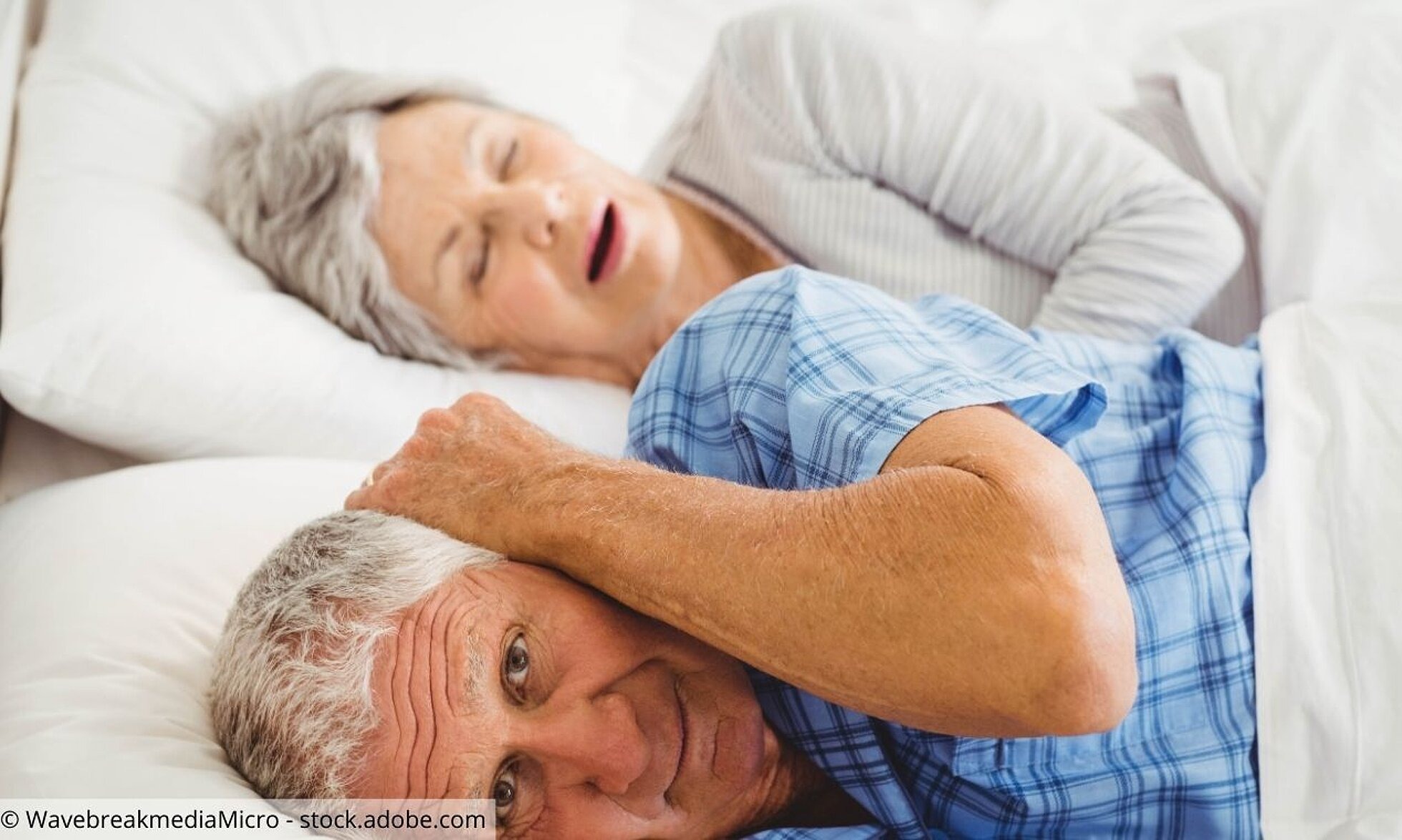 Älteres Ehepaar liegt zusammen im Bett, er hält sich die Ohren zu.