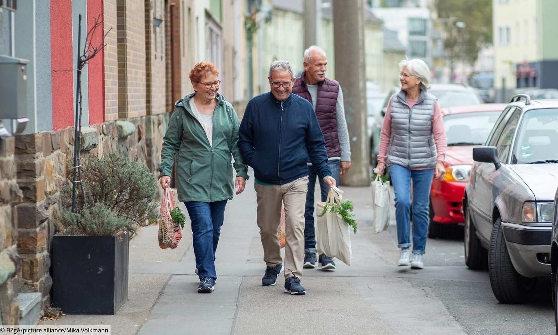 Vier ältere Erwachsener laufen gut gelaunt mit Einkaufstaschen einem Gehweg entlang