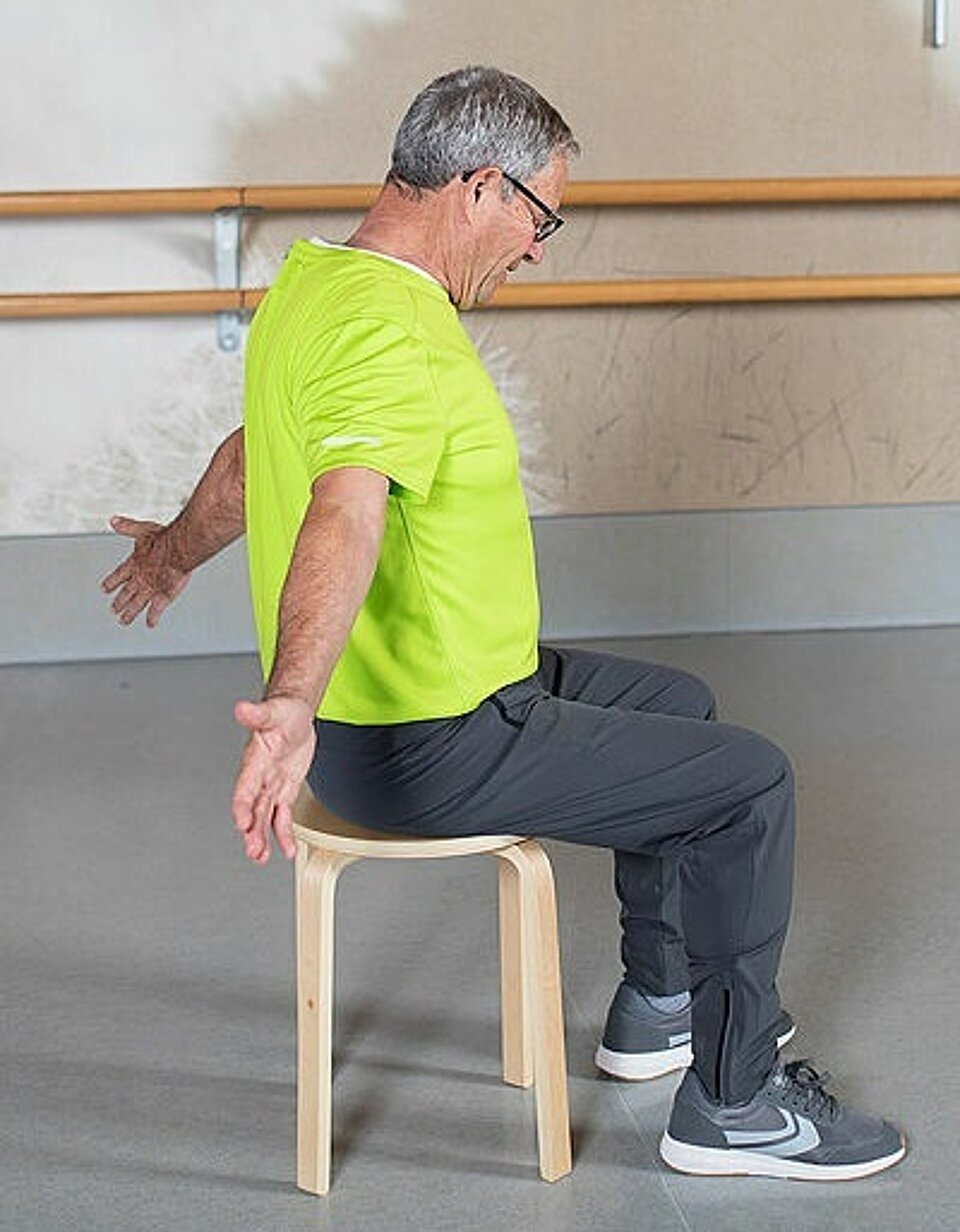 Mann sitzt auf Hocker und streckt beide Armen gerade nach hinten mit Daumen zur Decke.