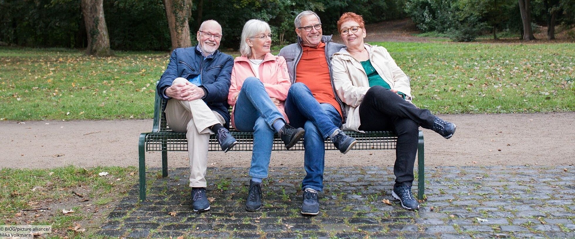 4 ältere Menschen sitzen auf einer Parkbank mit übereinandergeschlagenen Beinen.