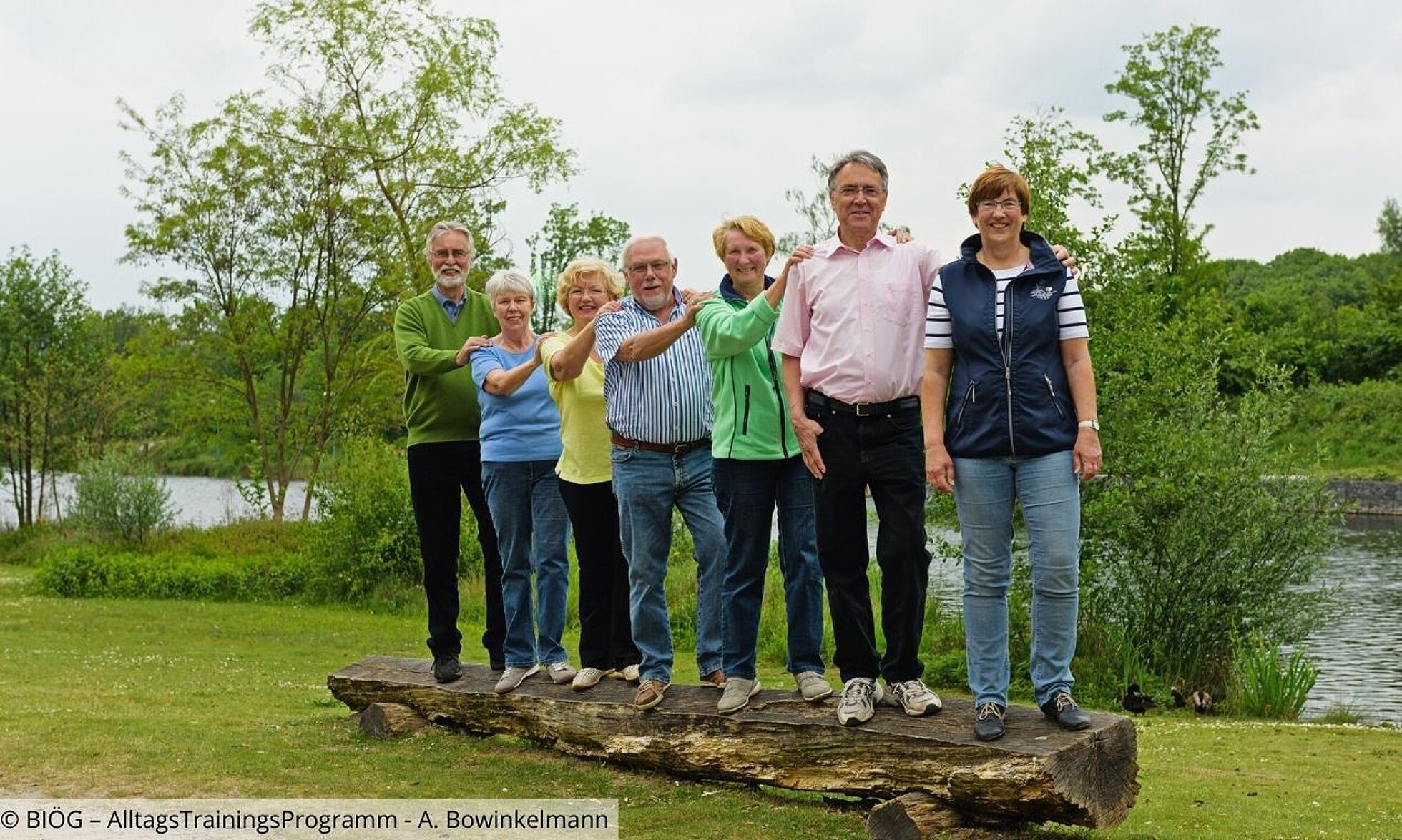 Gruppe von älteren Personen steht nebeneinander auf einen Baumstamm.