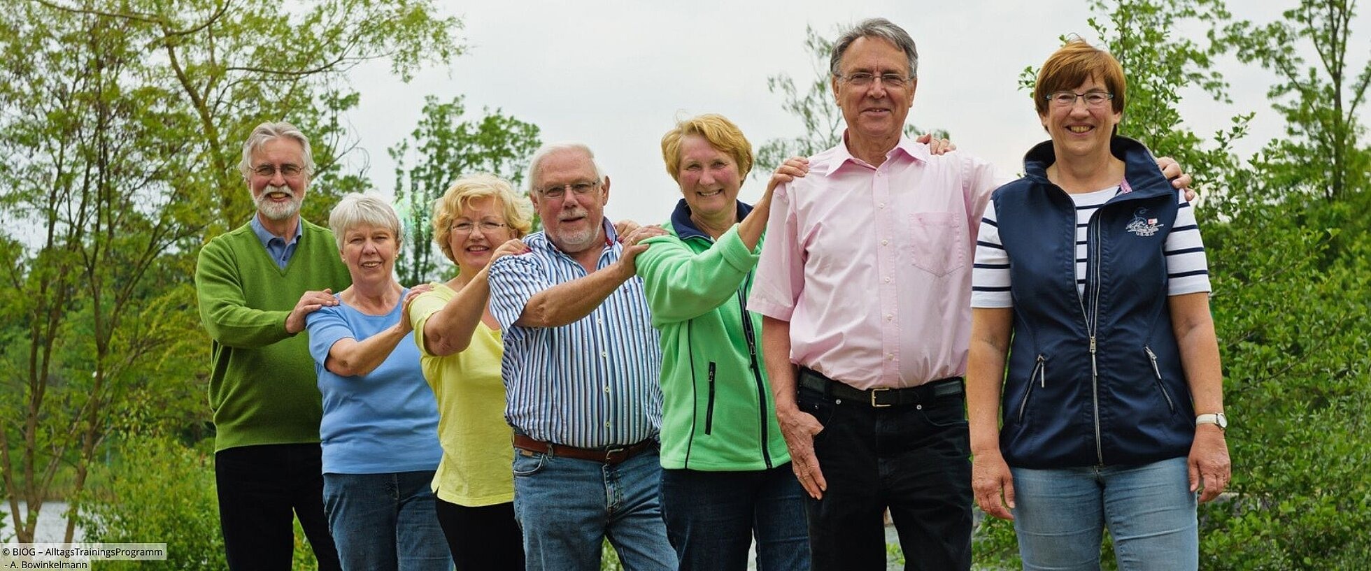 Gruppe von älteren Menschen steht hintereinander mit Händen auf den Schultern der vorderen Person.