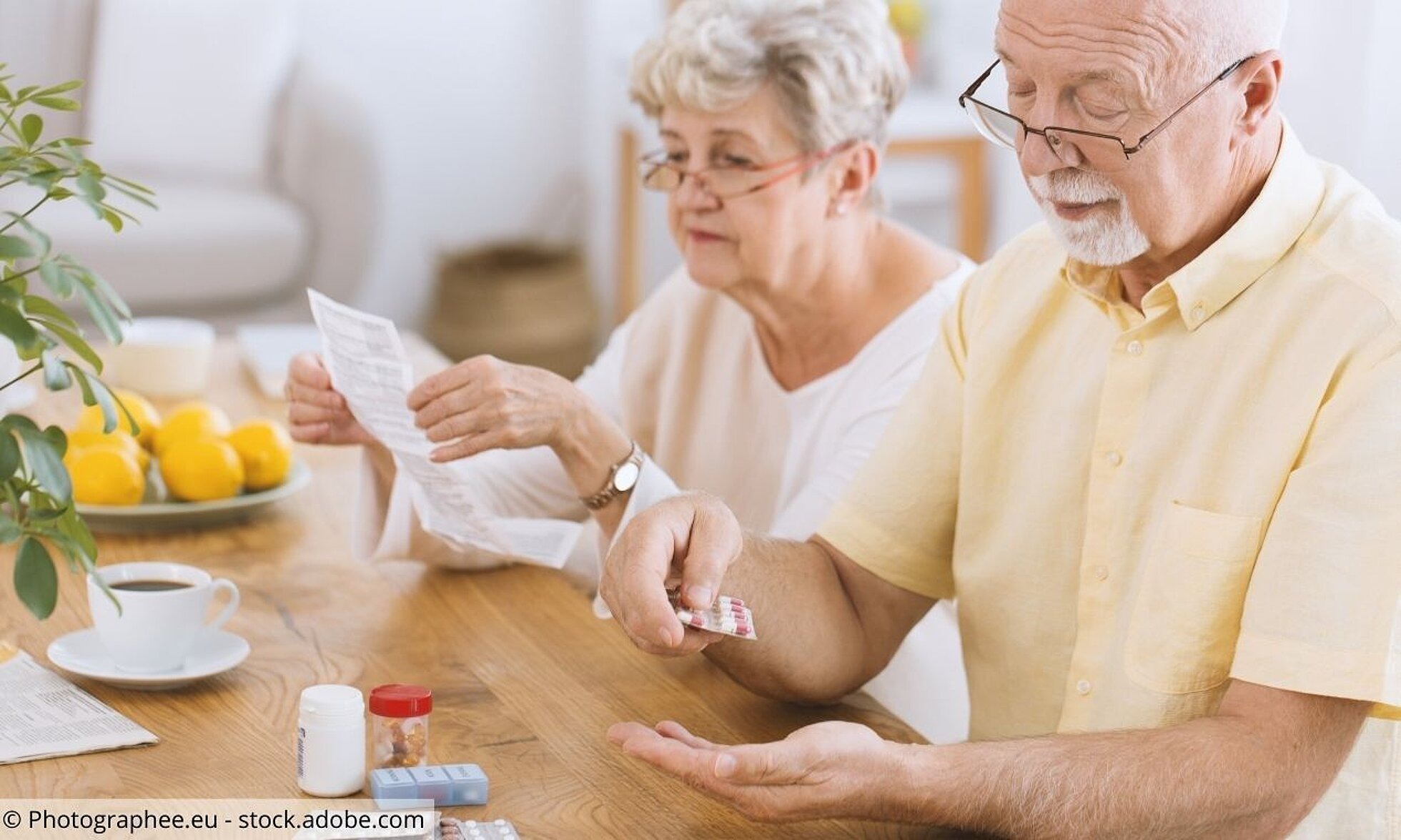 Älteres Paar am Frühstückstisch. Er nimmt Tabletten. Sie liest Beipackzettel.