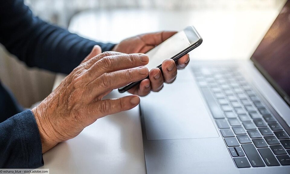 Senior tippt auf Smartphone mit Laptop im Hintergrund