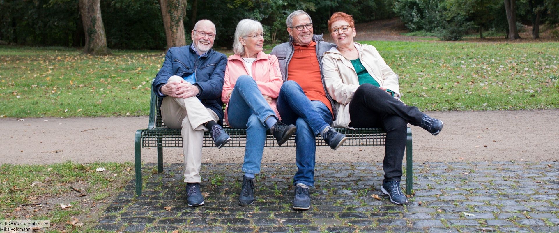 4 ältere Menschen sitzen auf einer Parkbank mit übereinandergeschlagenen Beinen.