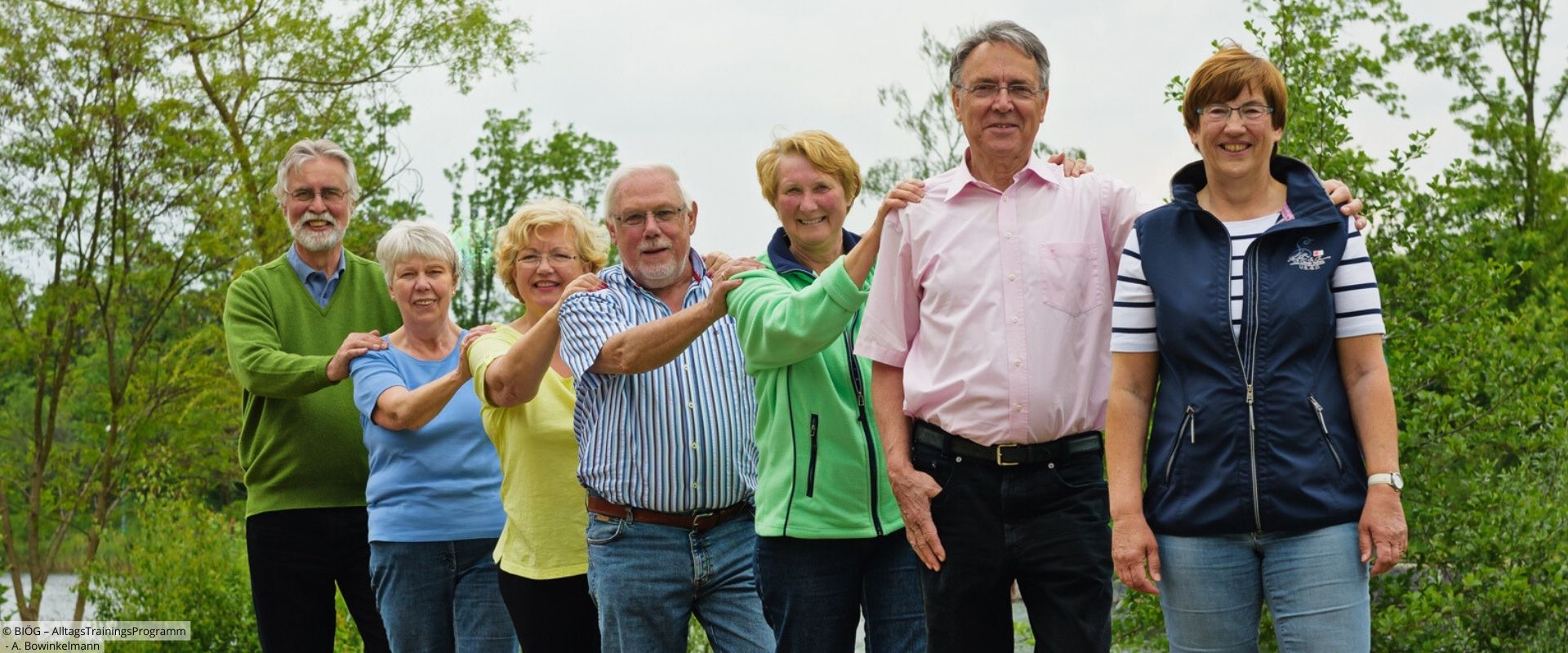 Gruppe von älteren Menschen steht hintereinander mit Händen auf den Schultern der vorderen Person.