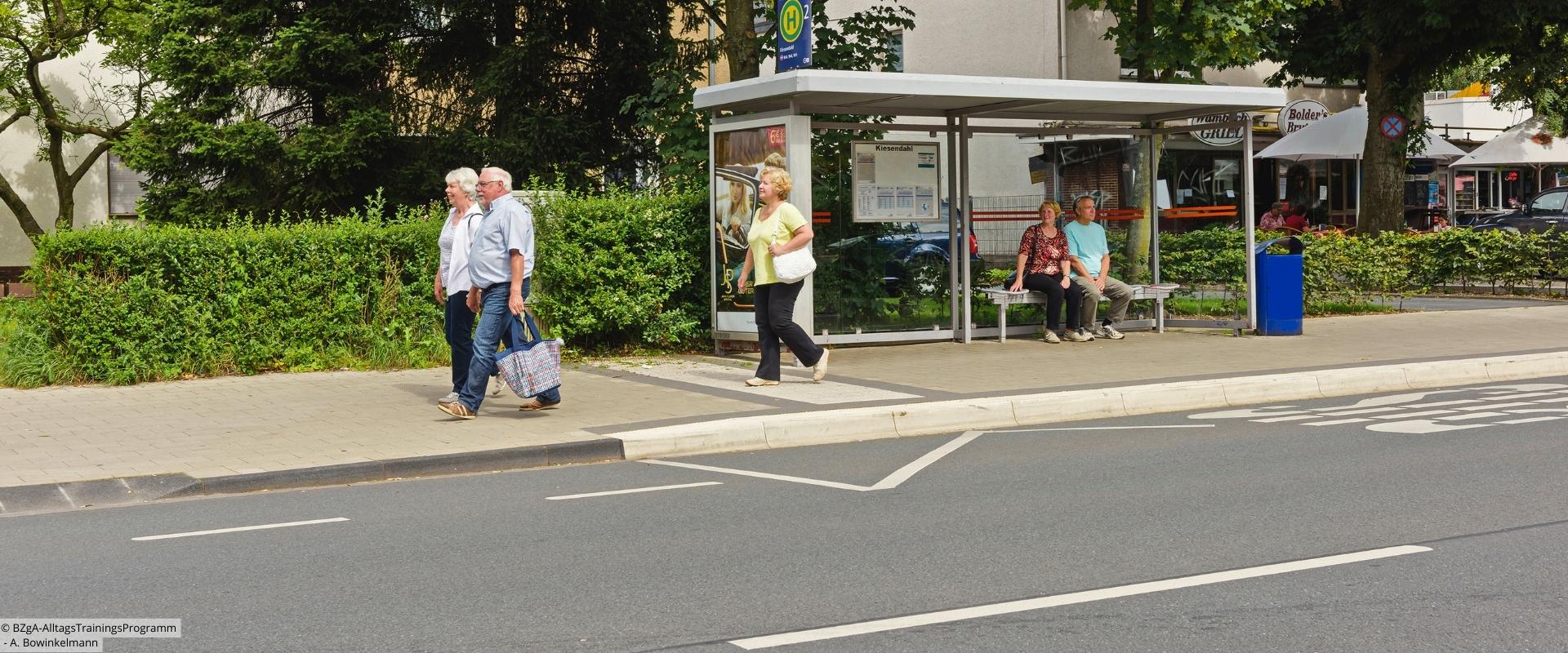 2 ältere Menschen sitzen an Bushaltestelle, 3 weitere laufen daran vorbei